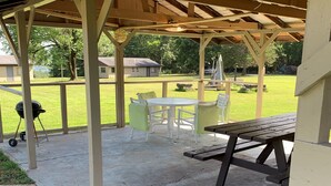 Een overdekt terras met een picknicktafel en stoelen, een barbecue en uitzicht op een gazon en huizen op de achtergrond.