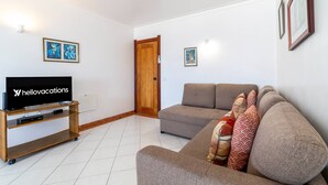 A living room with a flat-screen TV on a wooden stand, a grey sofa with patterned cushions, and two framed pictures on the wall. - Colibri Apartment by HelloVacations (Albufeira)