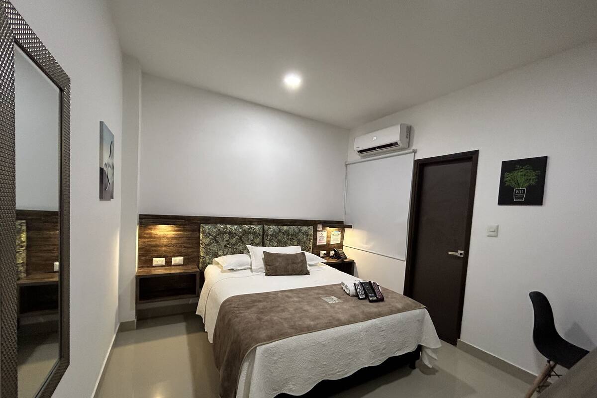 A hotel room with a bed, bedside table, mirror, wall-mounted air conditioner, and a door.