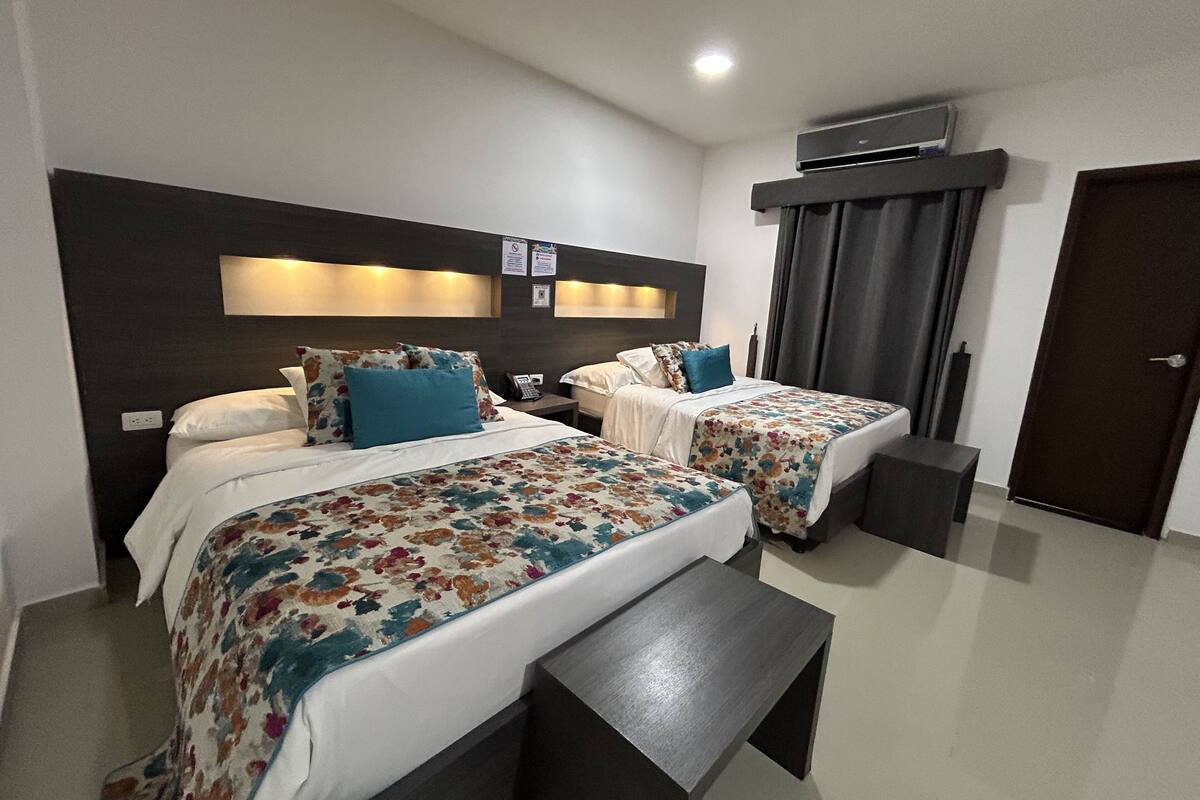 A hotel room with two beds, each with floral bedding and blue pillows. There is a bedside table with a phone and a small table in front of the beds. The room has a dark headboard, a ceiling light, and an air conditioning unit above the door.