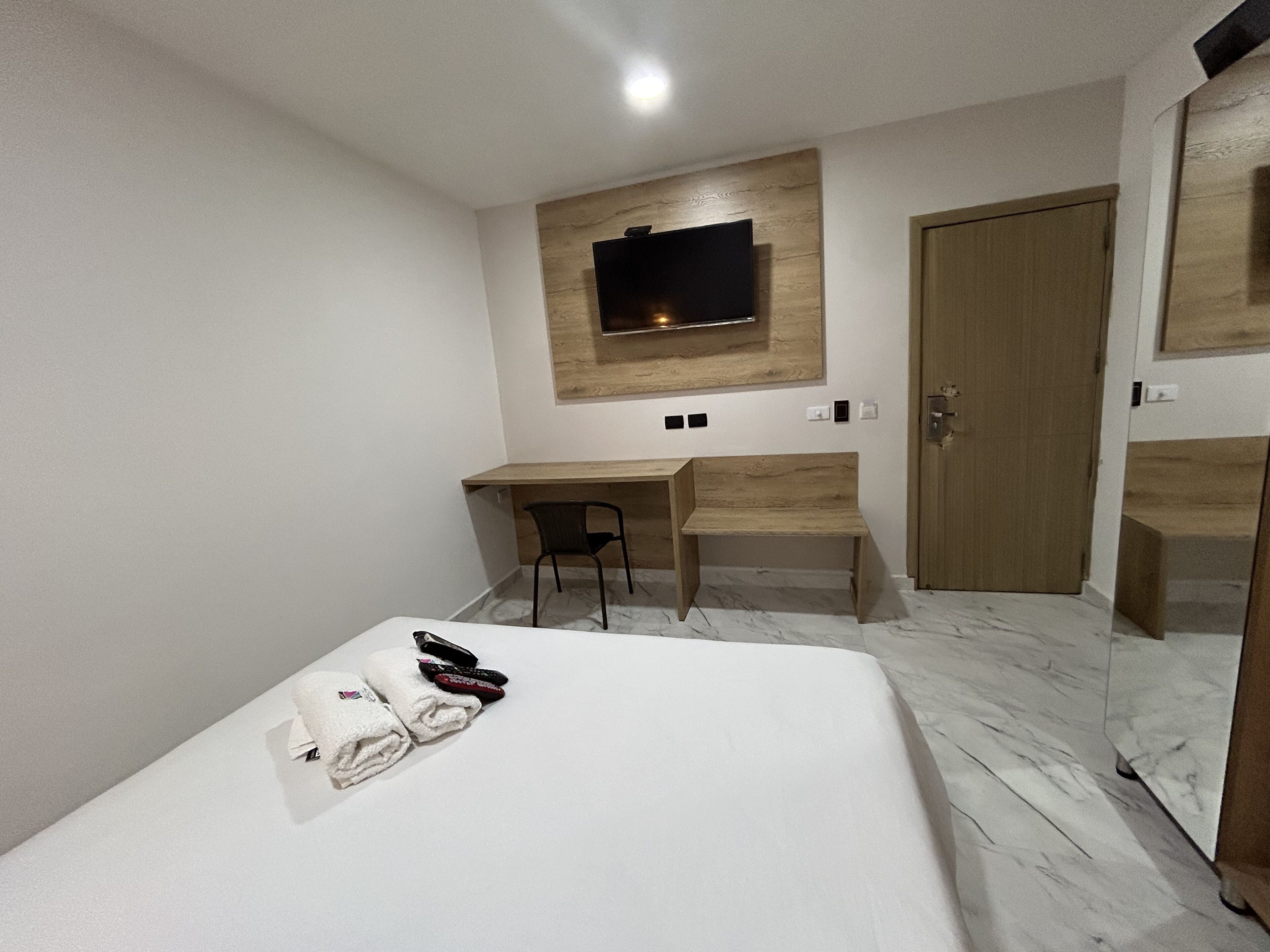 A hotel room with a bed, a desk, a chair, a TV mounted on the wall, and a wooden door.