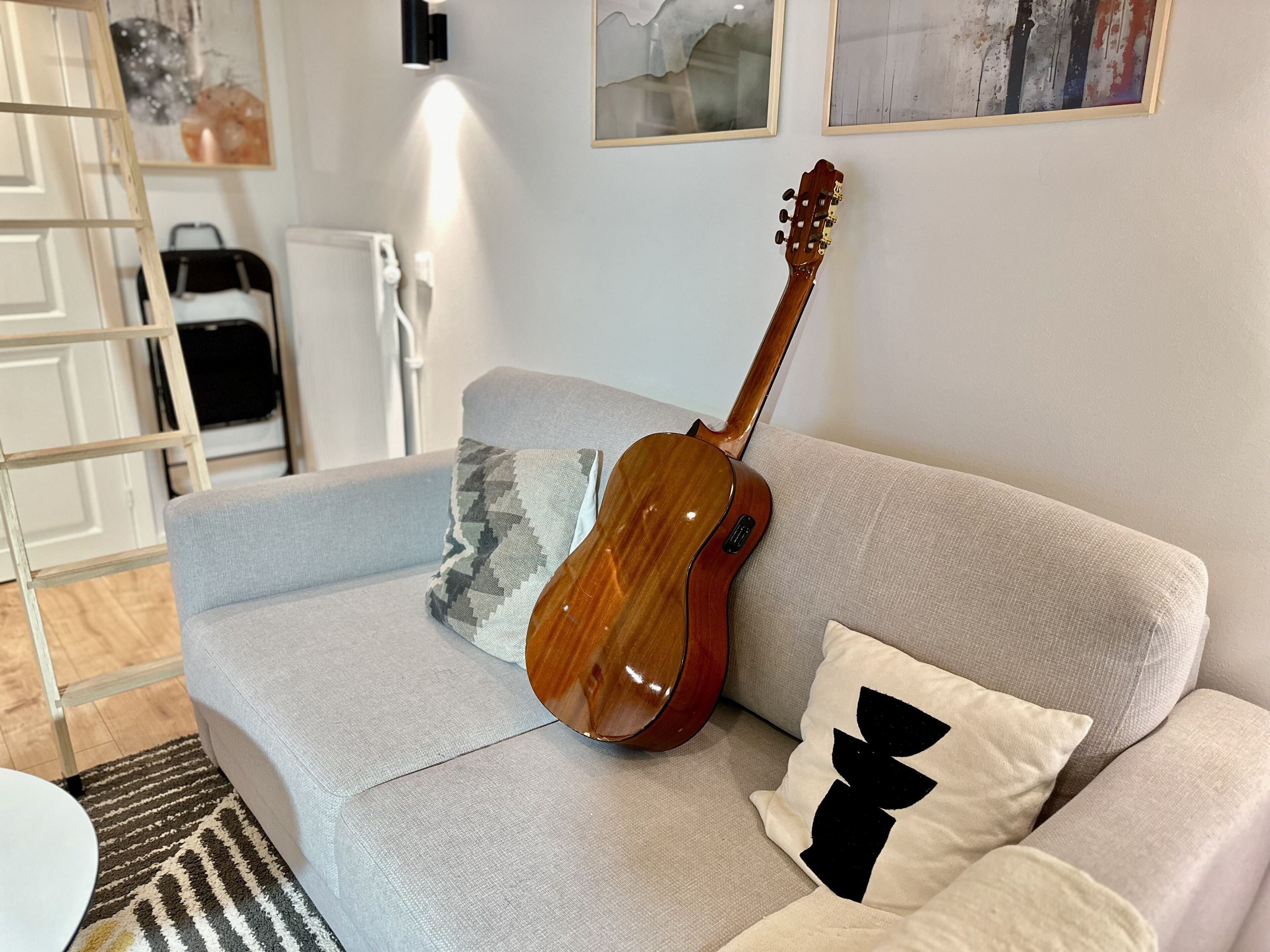 A modern living room with a grey sofa, a wooden guitar leaning against it, and a patterned throw pillow.