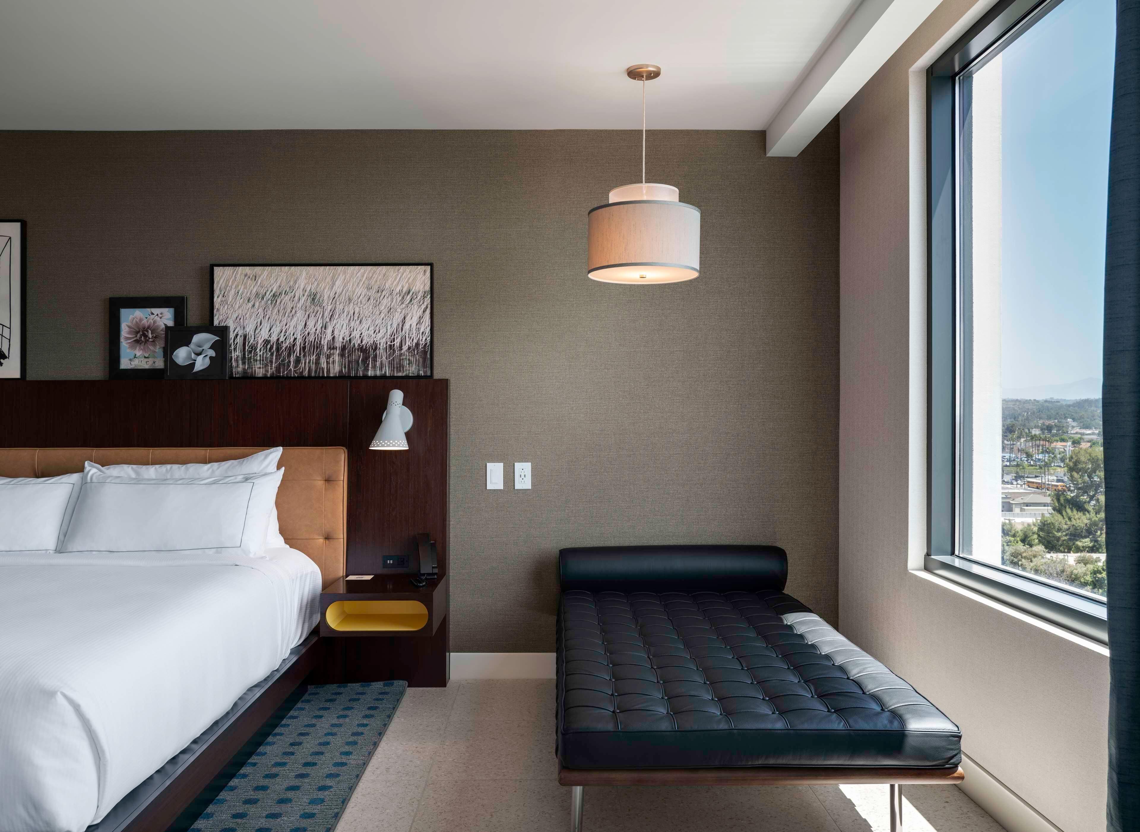 Una habitación de hotel moderna con una cama grande, un banco, una mesita de noche, una lámpara de pared y vistas a la ciudad.