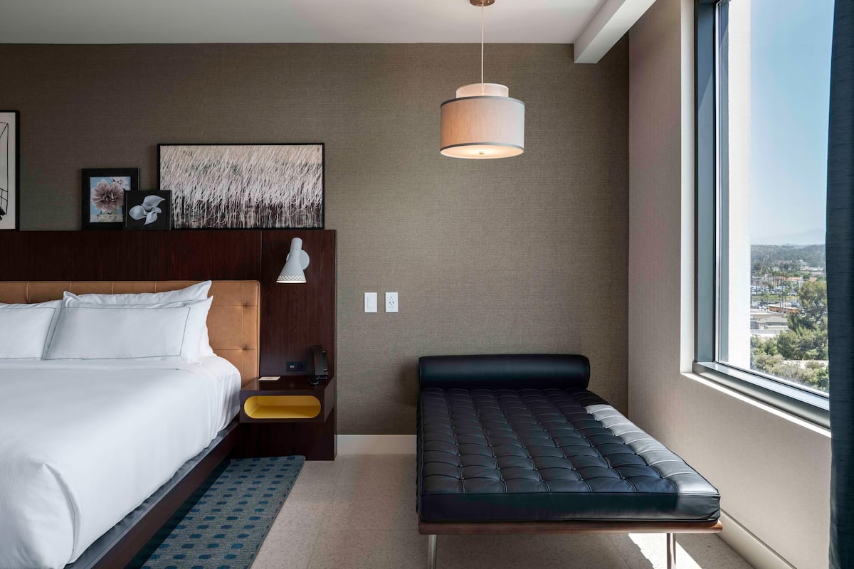 Una habitación de hotel moderna con una cama grande, un banco, una mesita de noche, una lámpara de pared y vistas a la ciudad.