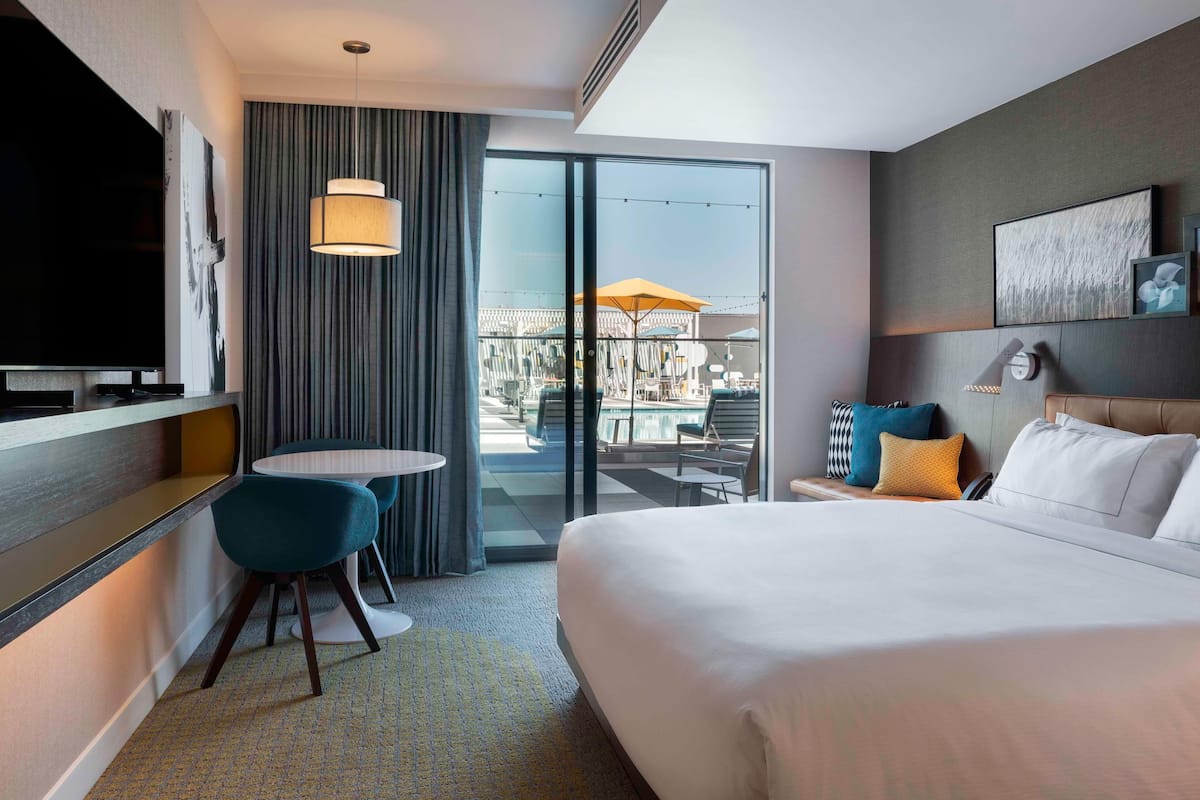 Habitación de hotel con una cama grande, un escritorio con silla, televisor, balcón con asientos exteriores y puerta corrediza de vidrio.