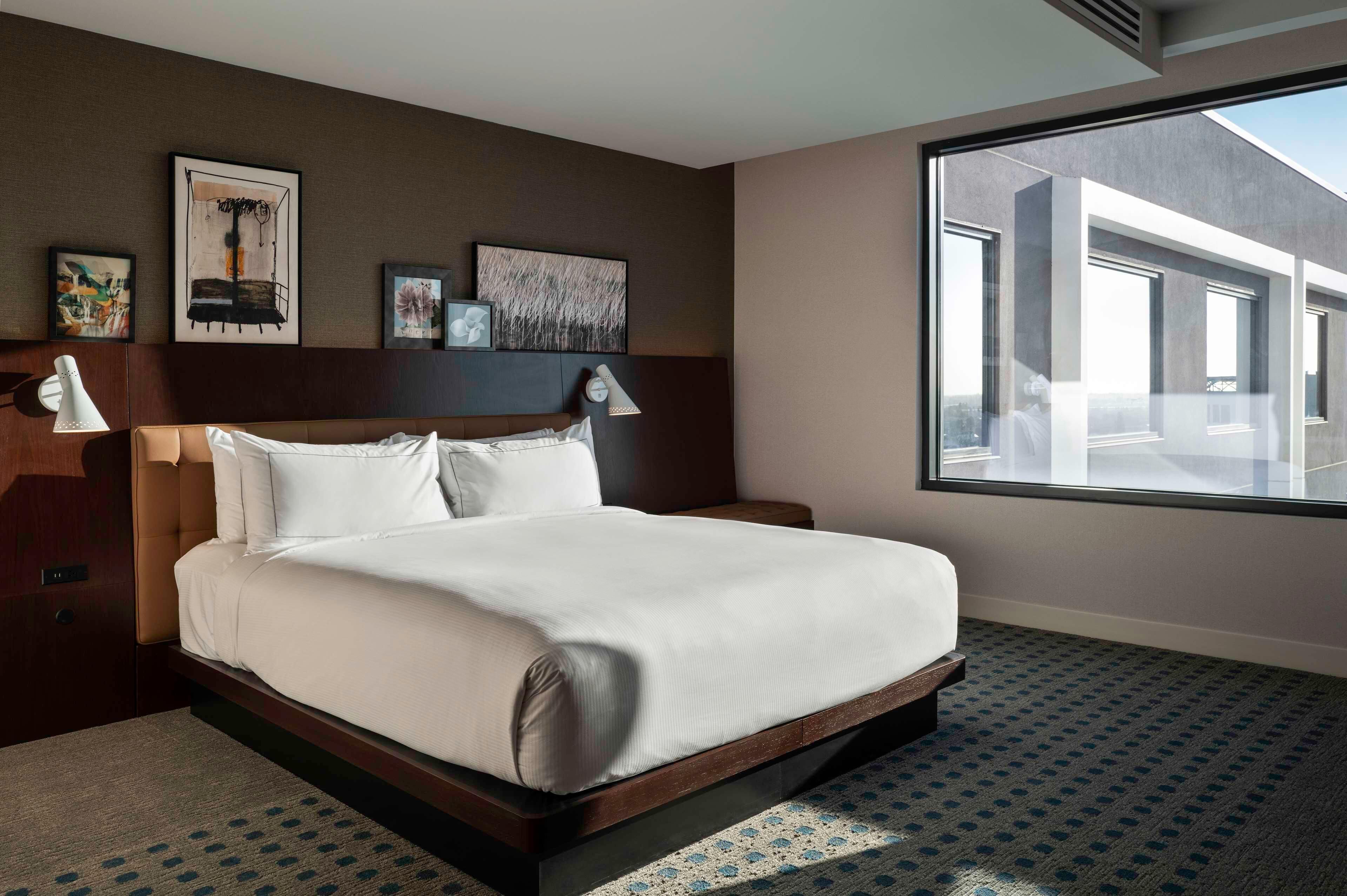 Habitación de hotel moderna con una cama grande, dos lámparas de noche, un cabecero y un ventanal con vistas a la ciudad.