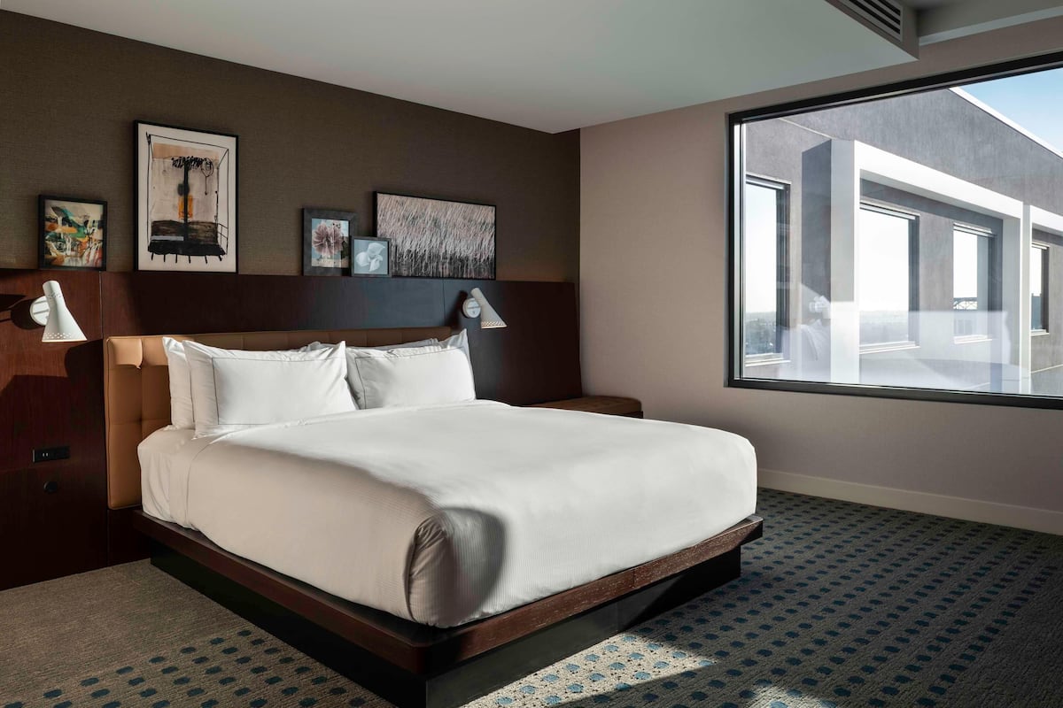 Habitación de hotel moderna con una cama grande, dos lámparas de noche, un cabecero y un ventanal con vistas a la ciudad.