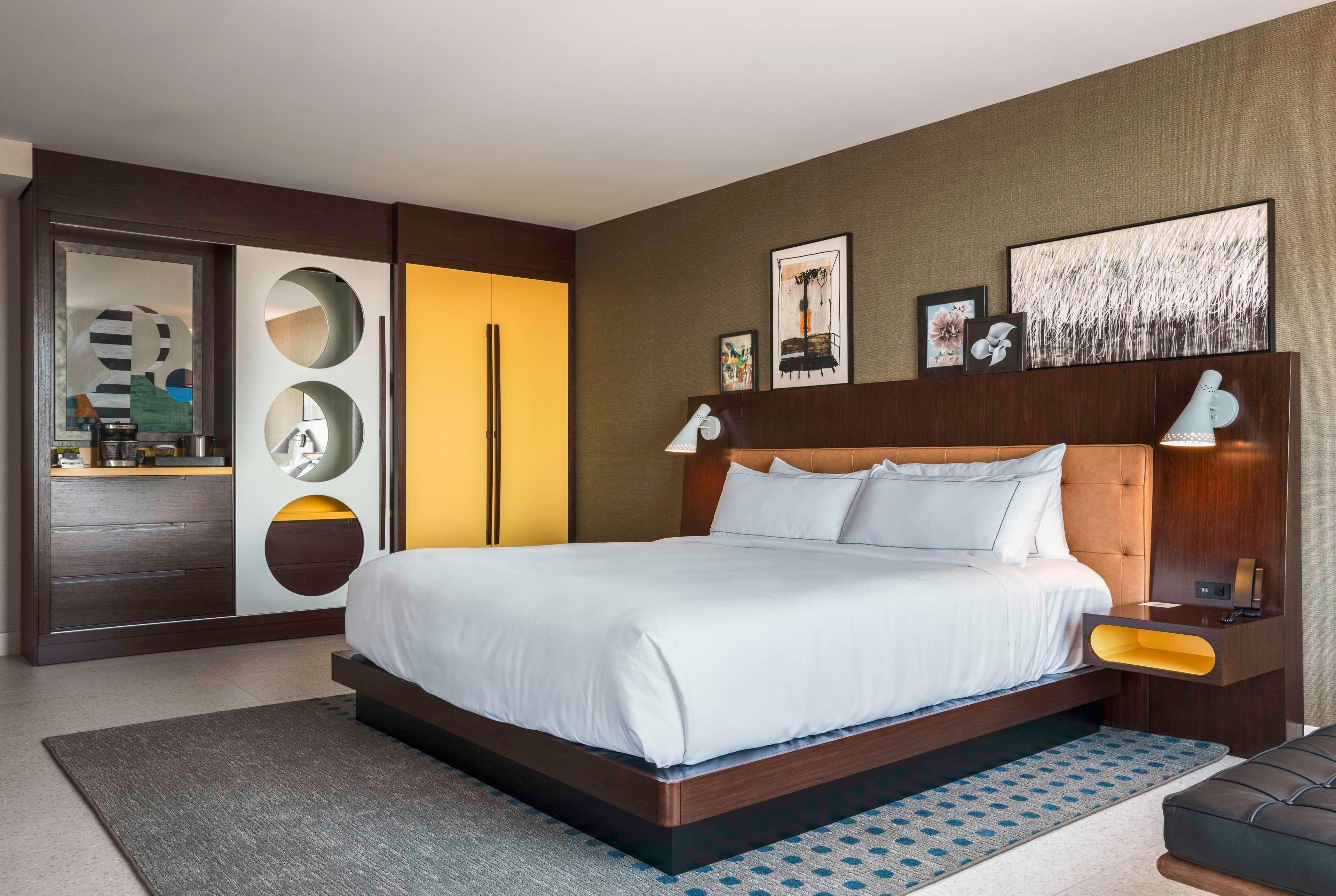 Una habitación de hotel moderna con una cama grande, un armario con espejos circulares, una mesita de noche con lámpara y obras de arte en las paredes.