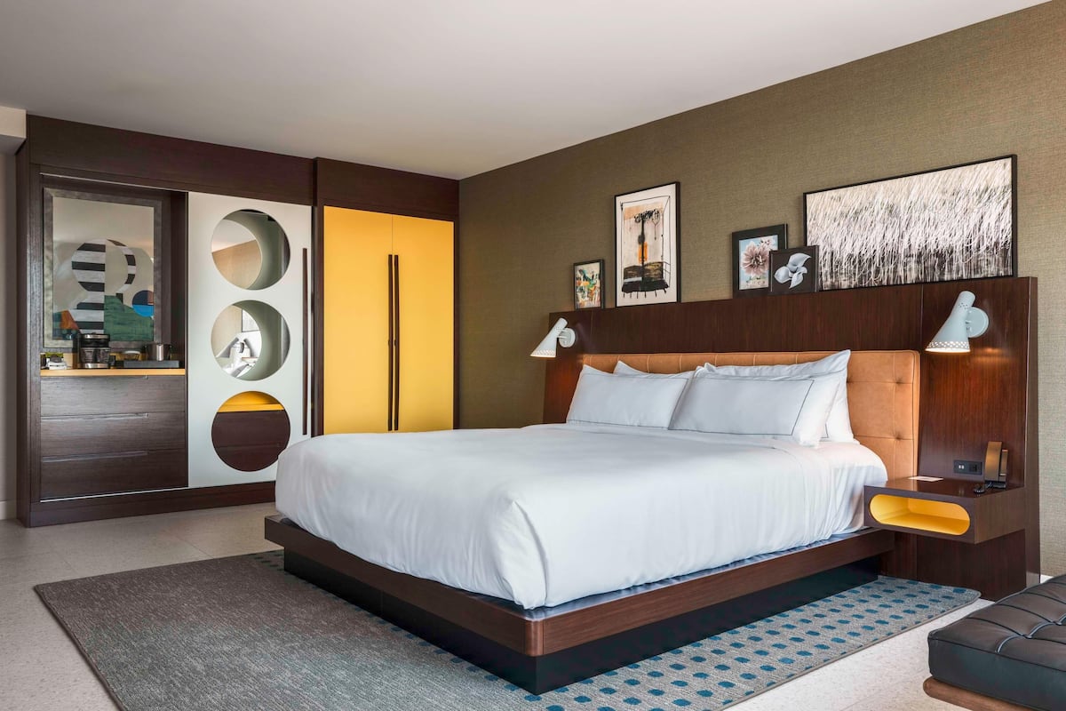 Una habitación de hotel moderna con una cama grande, un armario con espejos circulares, una mesita de noche con lámpara y obras de arte en las paredes.