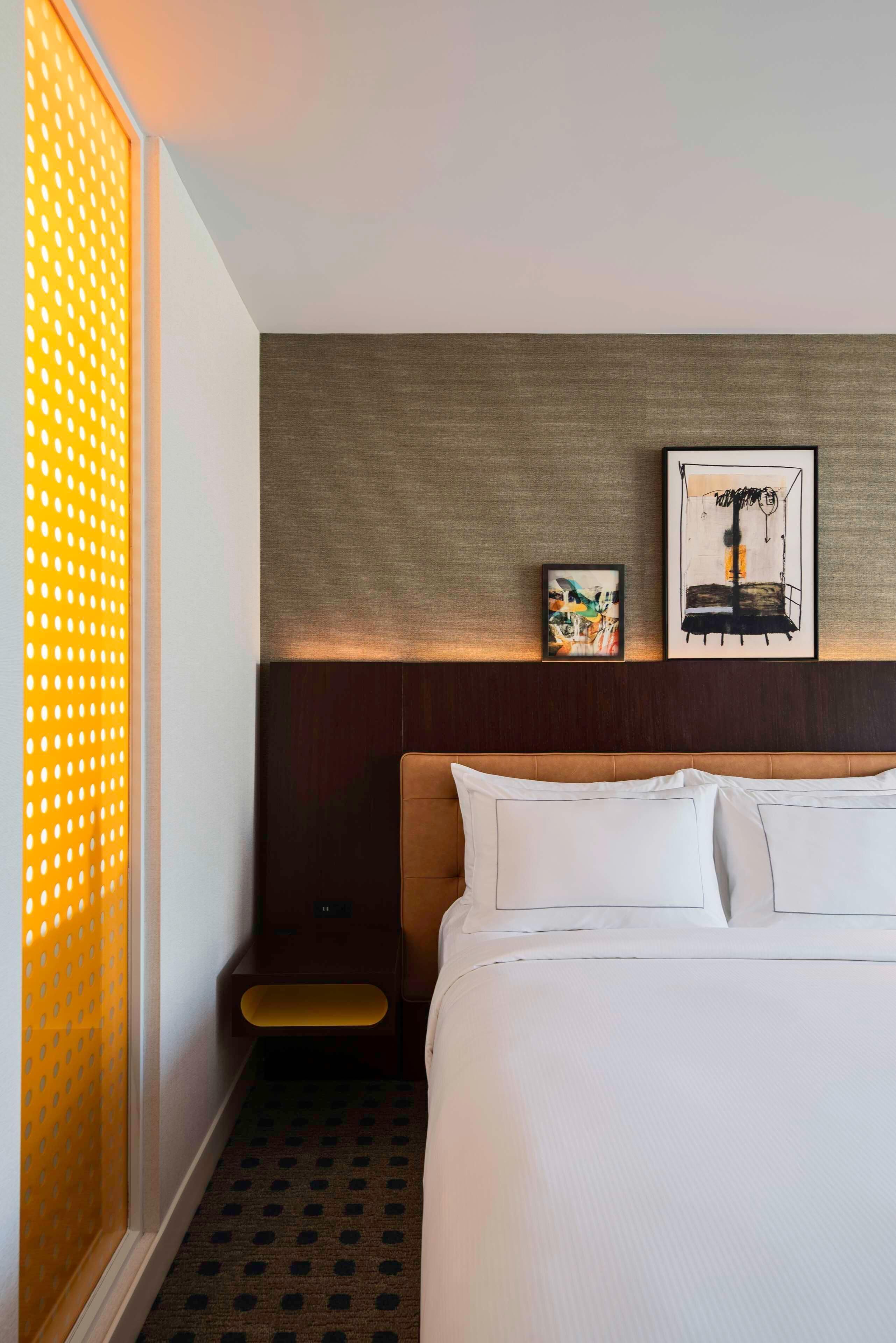 Habitación de hotel con una cama grande, cabecero con pared texturizada, mesita de noche pequeña y dos obras de arte enmarcadas en la pared.