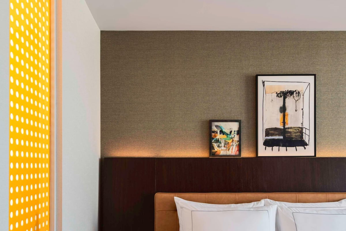 Habitación de hotel con una cama grande, cabecero con pared texturizada, mesita de noche pequeña y dos obras de arte enmarcadas en la pared.