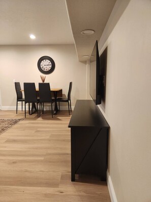 Ein Essbereich mit einem Holztisch, vier Stühlen, einer Wanduhr und einem flachbildfernseher an der Wand.