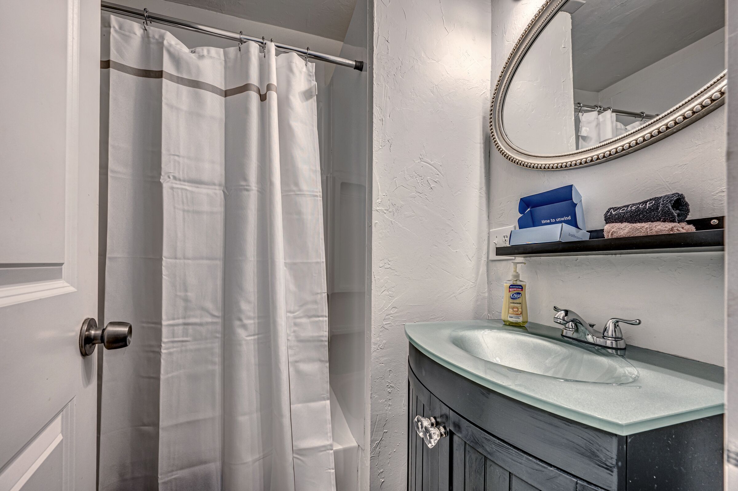 A bathroom with a glass sink, a mirror, a shower curtain, and toiletries on a shelf.