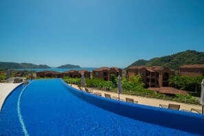 A resort with a long swimming pool, villas with red-tiled roofs, and a view of the sea and hills.