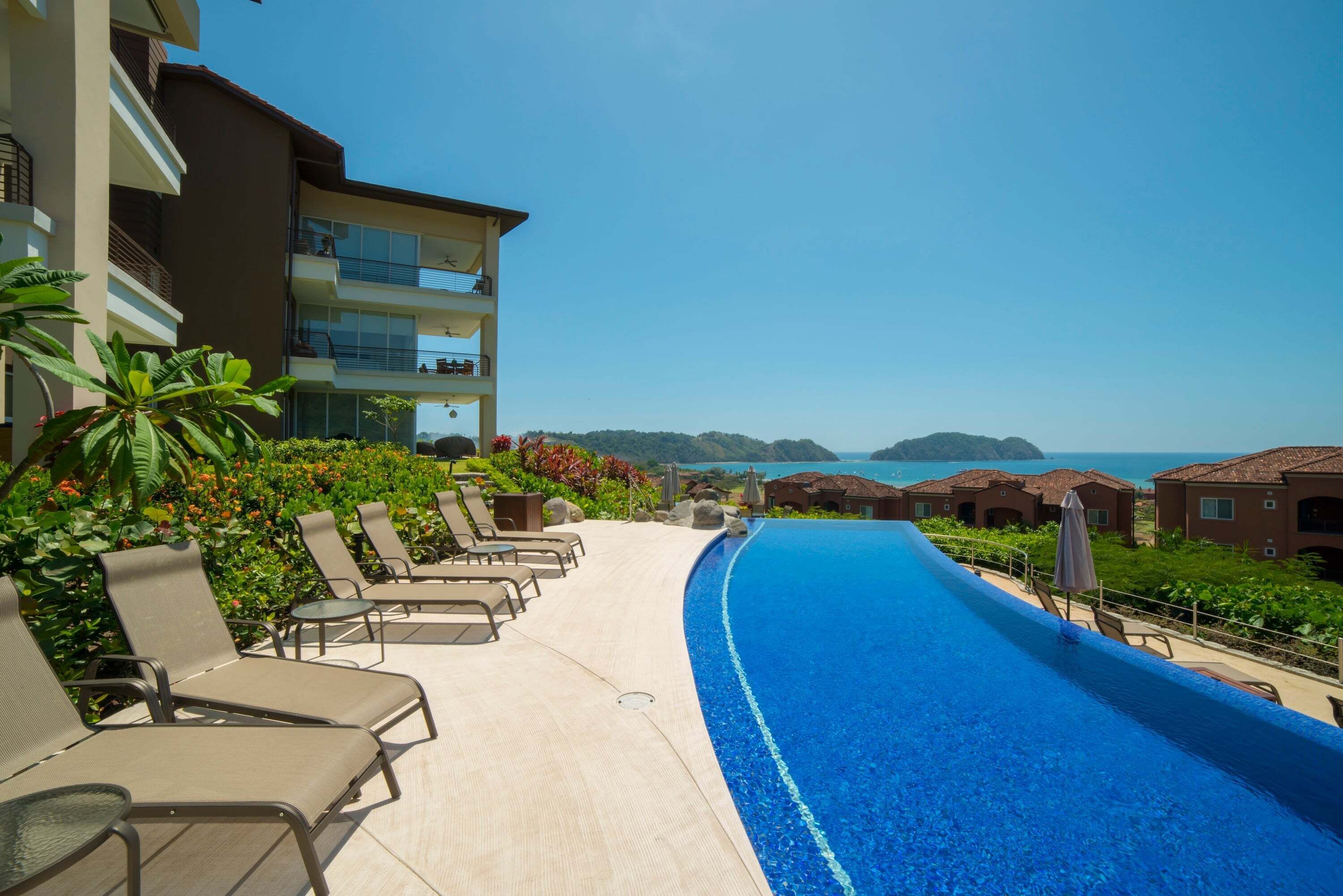 Zona de piscina en un resort con sillones de descanso, una piscina azul cristalina y vistas al mar y las colinas.