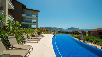 Zona de piscina en un resort con sillones de descanso, una piscina azul cristalina y vistas al mar y las colinas.