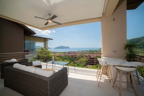 A balcony with a wicker sofa, a glass table, and a bar stool.