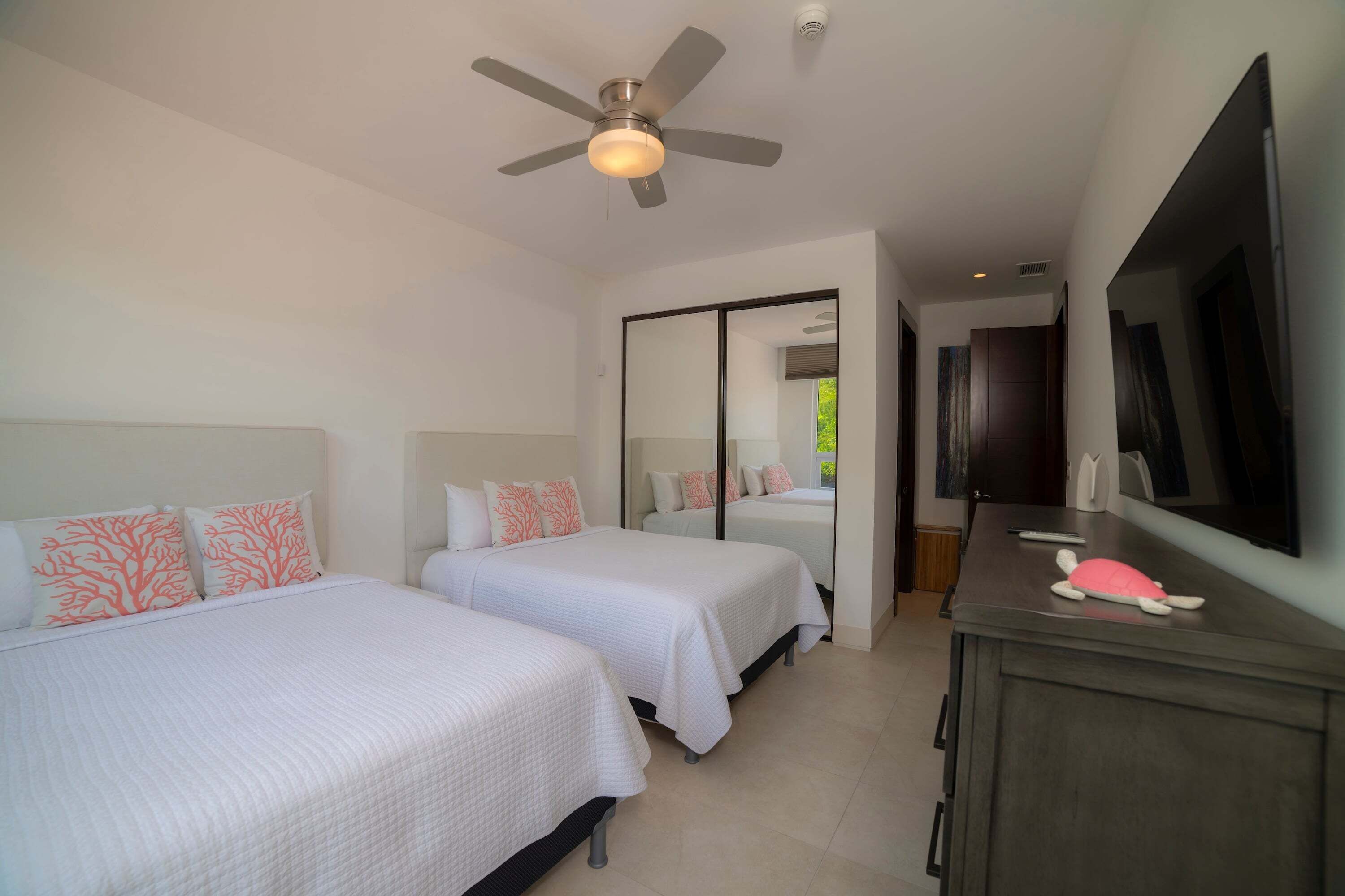 Habitación de hotel con dos camas, un ventilador de techo, un espejo grande, un televisor y una mesita con un control remoto.