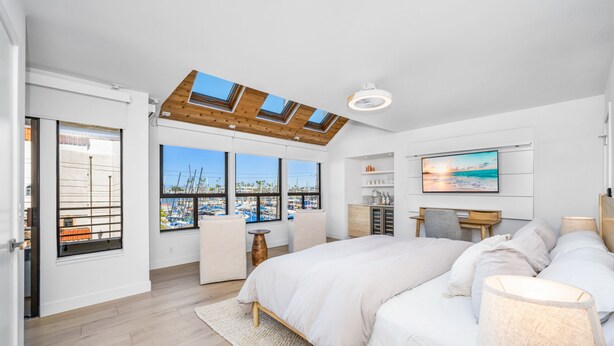 A bedroom with a large bed, a desk, a chair, a TV, and a view of boats through large windows.