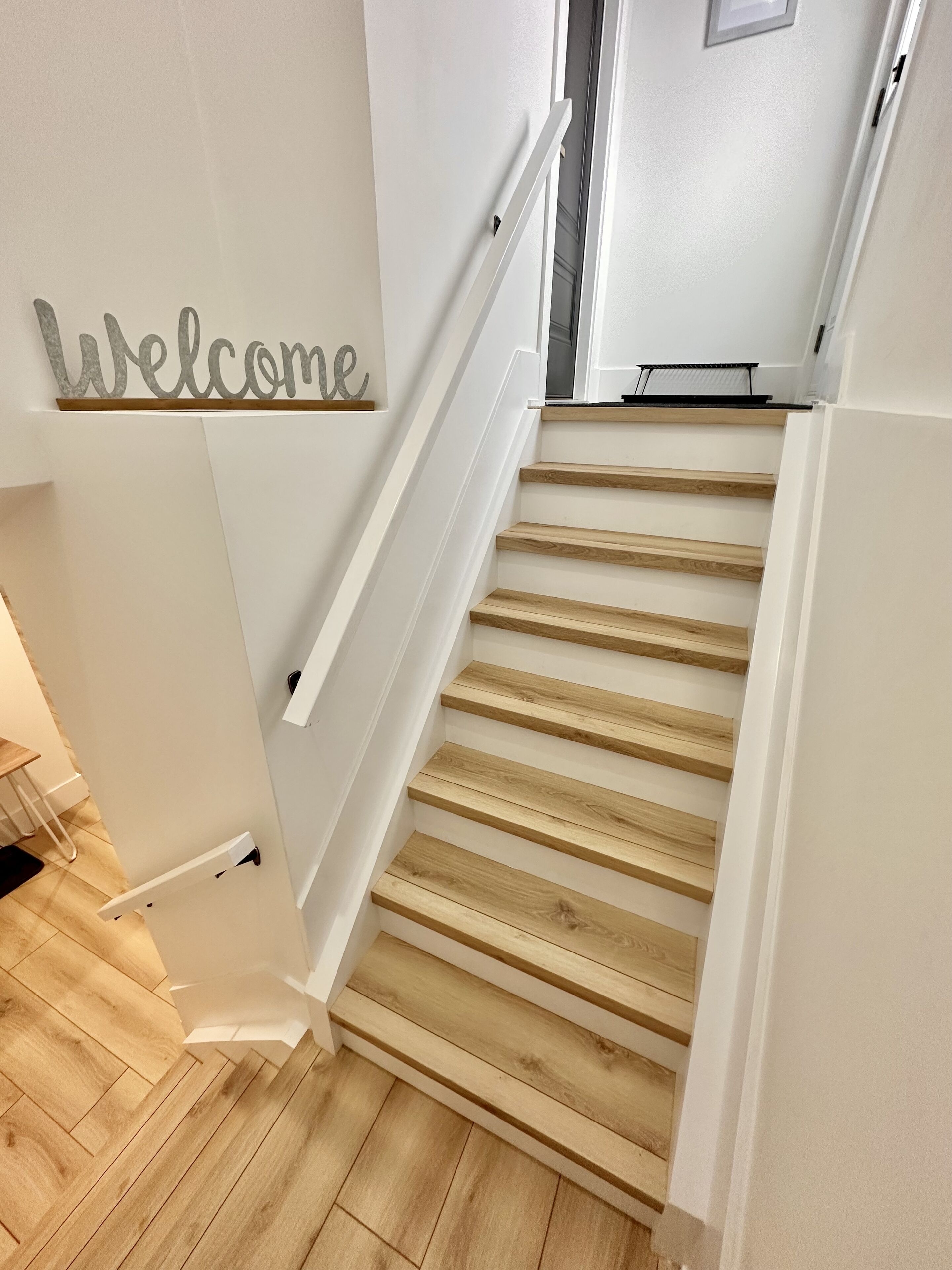 Un escalier avec des marches en bois et des murs blancs, menant à une porte sur laquelle est affiché un panneau indiquant « Bienvenue ».