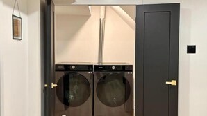 A laundry room with two front-loading washing machines, a door, and a white wall with a framed picture. - Modern, Bright, Spacious 2 Bedroom Guest Suite - Entire Separate Basement  (Grimsby)
