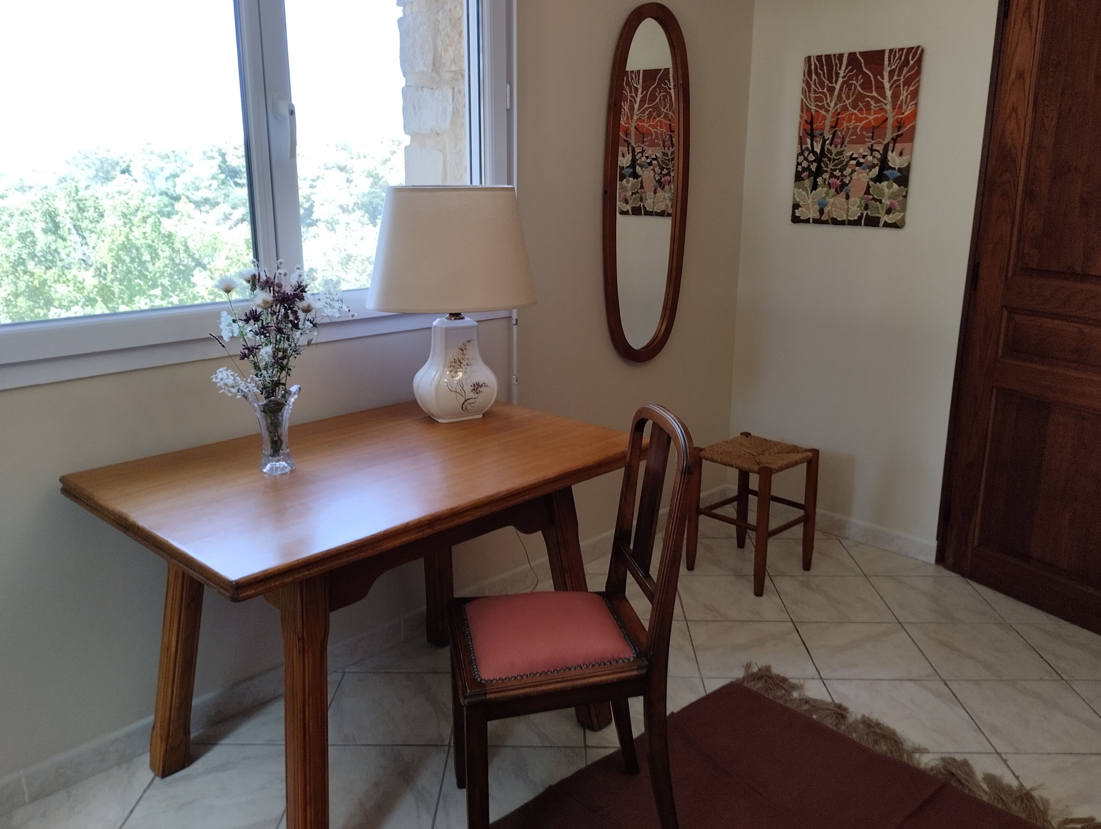 A wooden dining table with a vase of flowers, a chair with a red cushion, a small stool, a mirror, and two wall art pieces.