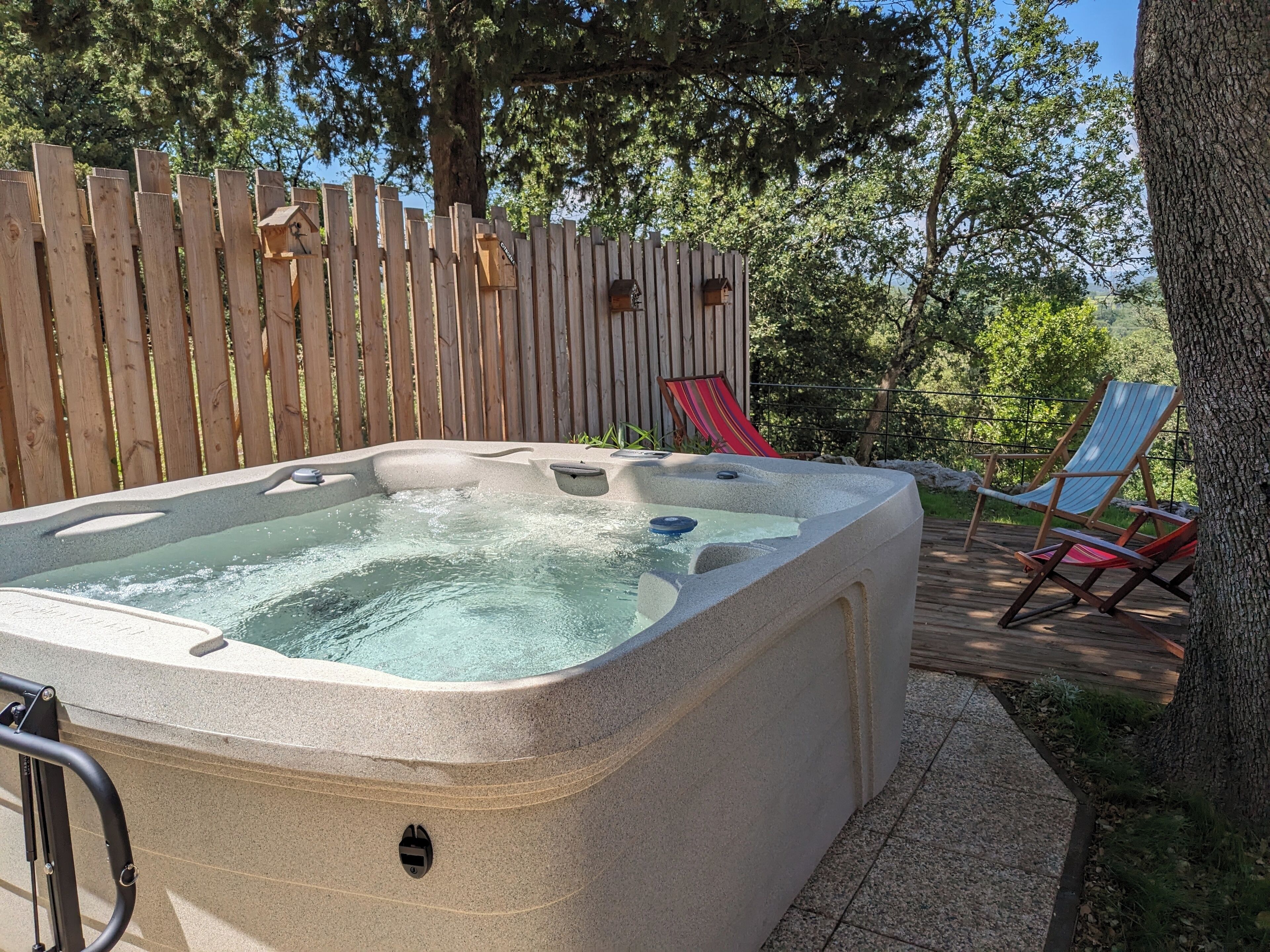A hot tub with a wooden deck and chairs, surrounded by trees.