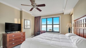 Habitación de hotel con una cama grande, un ventilador de techo, un televisor sobre un aparador de madera, una ventana con vista al océano y una lámpara en una mesita de noche.