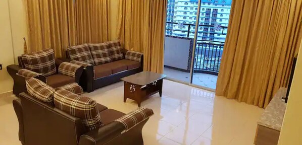 A living room with a sofa set and a coffee table, large windows with curtains, and a view of a high-rise building.