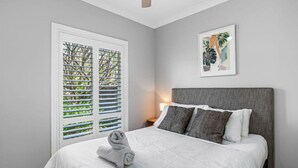 A bedroom with a bed, a nightstand, a lamp, a window with blinds, and a painting on the wall.