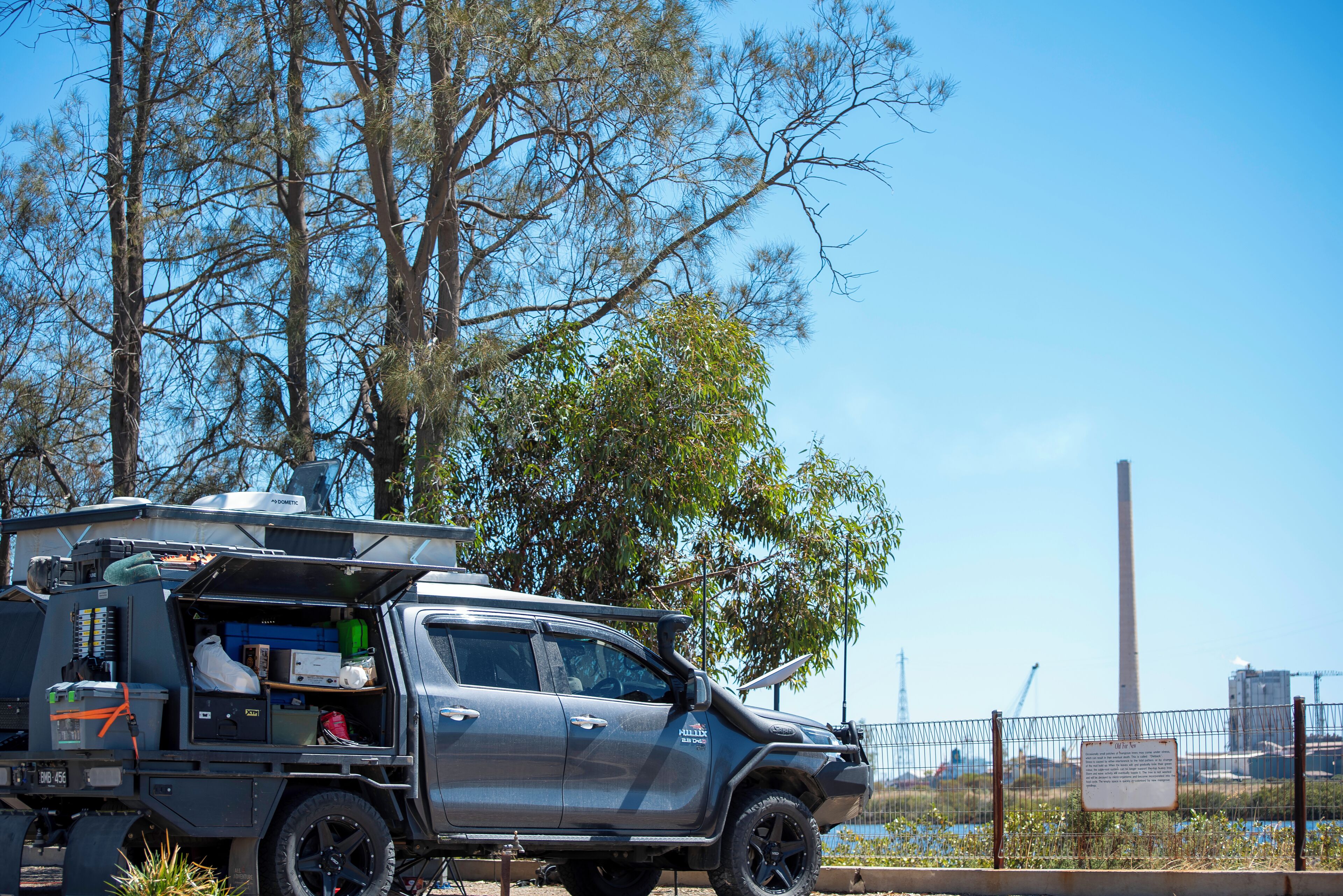 A silver pickup truck with an open cargo bed, parked near a fence with trees and a building in the background.