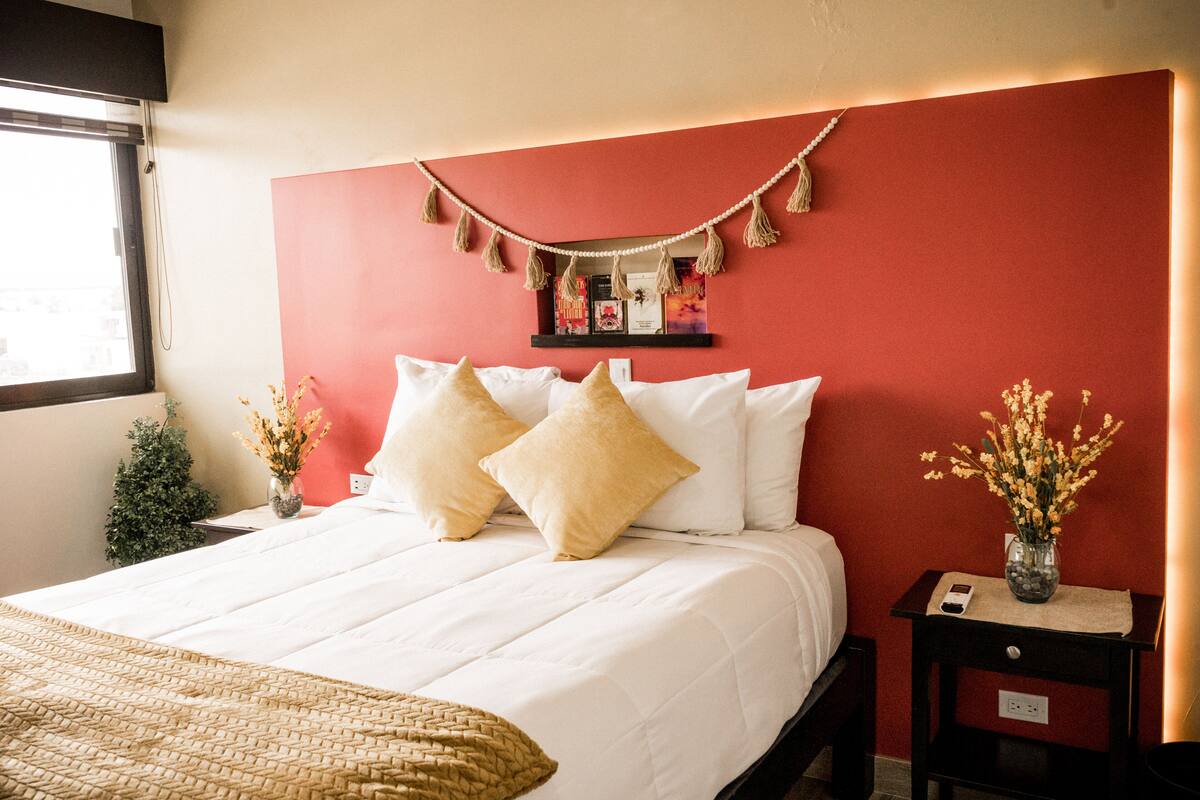 Un lit bien fait, avec une literie blanche et beige, un mur d’accent rouge et une petite table de chevet ornée d’un vase de fleurs.