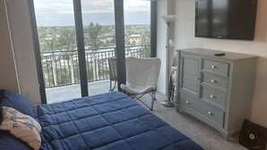 Une chambre à coucher comprenant un lit, une chaise, une commode, un téléviseur et un balcon avec vue sur la ville.
