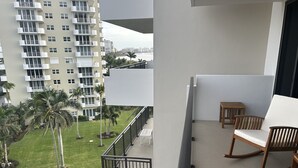 Un balcon avec deux fauteuils à bascule en bois et une petite table, donnant sur un espace verdoyant agrémenté de palmiers et sur un bâtiment en arrière-plan.