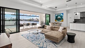 A modern living room with a large sectional sofa, a dining area with a view of a body of water, and a kitchen with white cabinets.