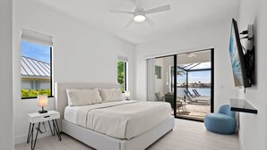 A modern bedroom with a large bed, a nightstand, a desk, a chair, and a view of the outdoors through a sliding glass door.