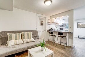 A modern living room with a grey sofa, striped cushions, a white coffee table with a plant, and a kitchen area with a bar stool and a microwave.