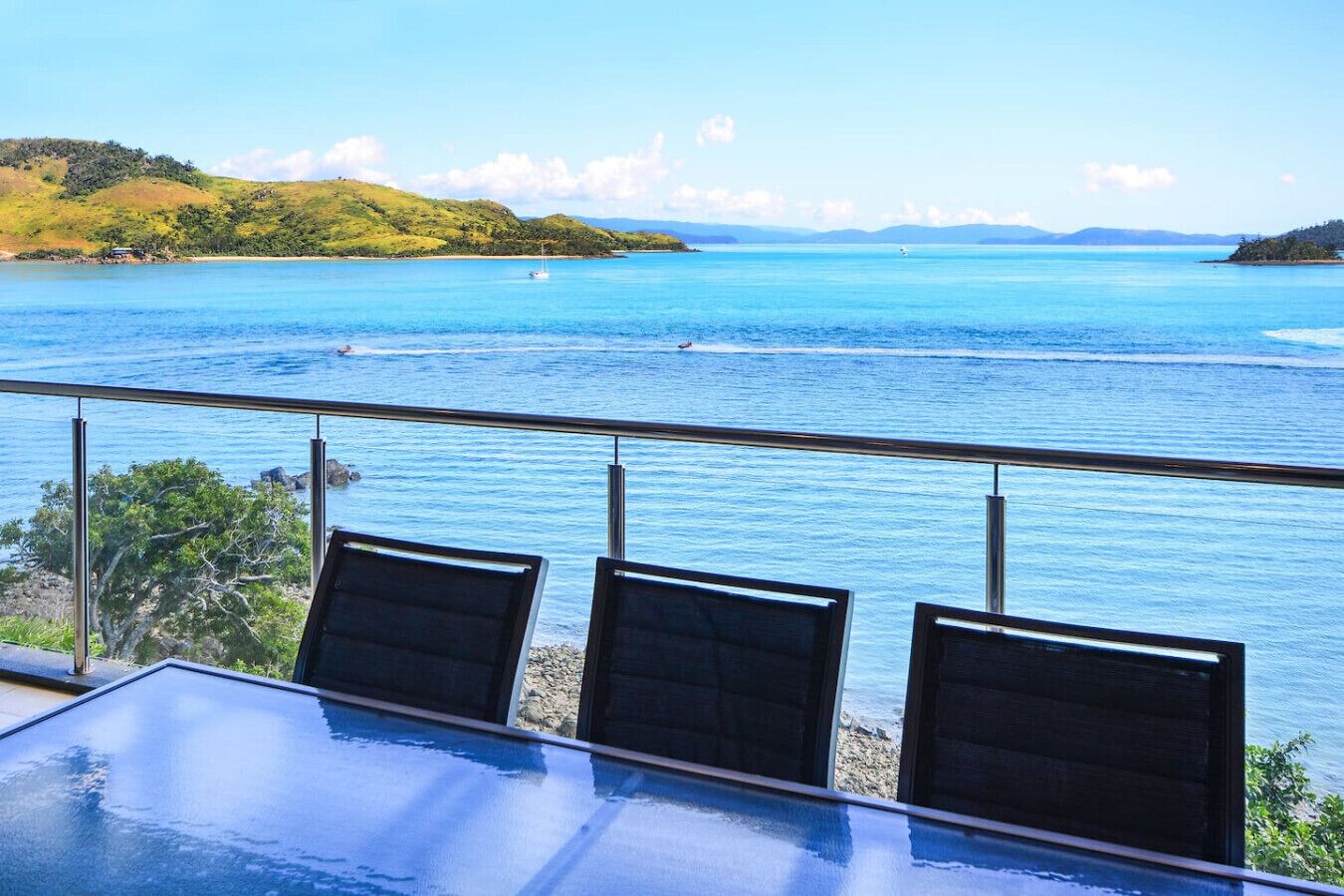 Un balcon avec une table et des chaises en verre, donnant sur une vaste étendue d’eau, avec des collines à l’horizon.