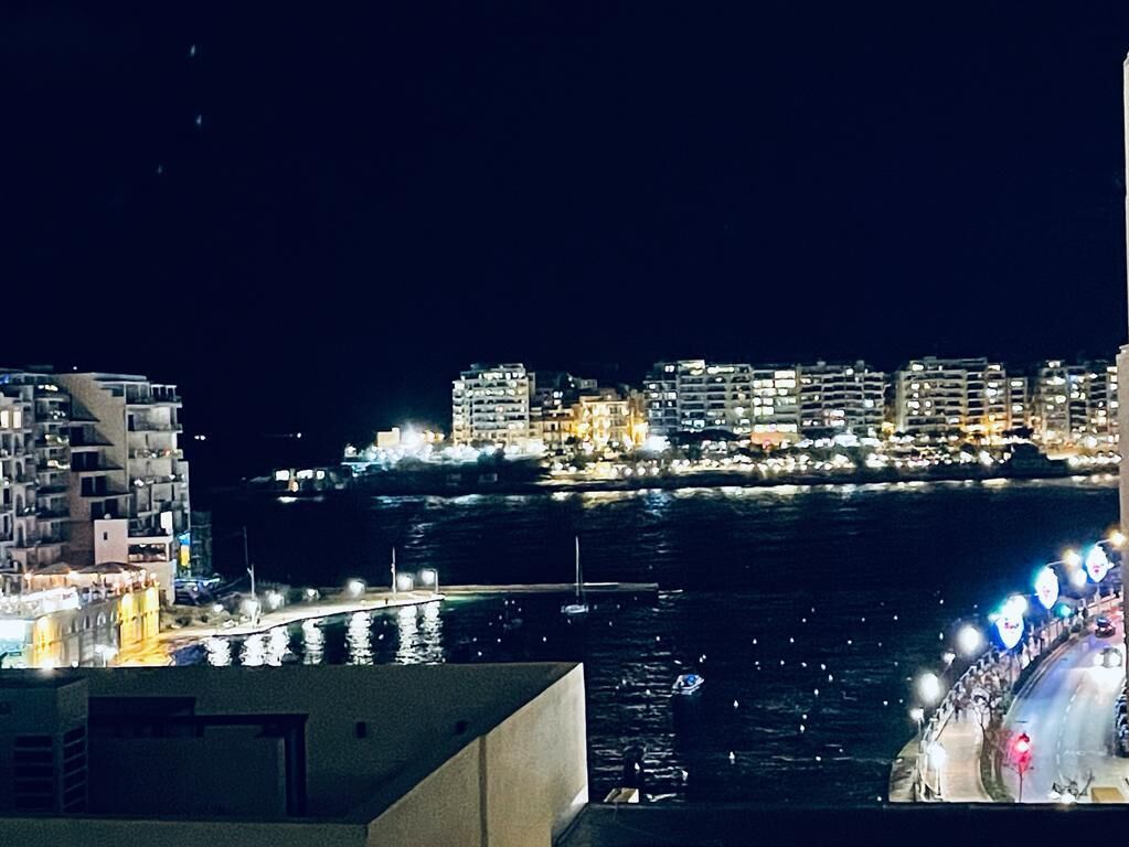 Nachtansicht einer Stadt mit Uferpromenade, beleuchteten Gebäuden und einer Brücke.