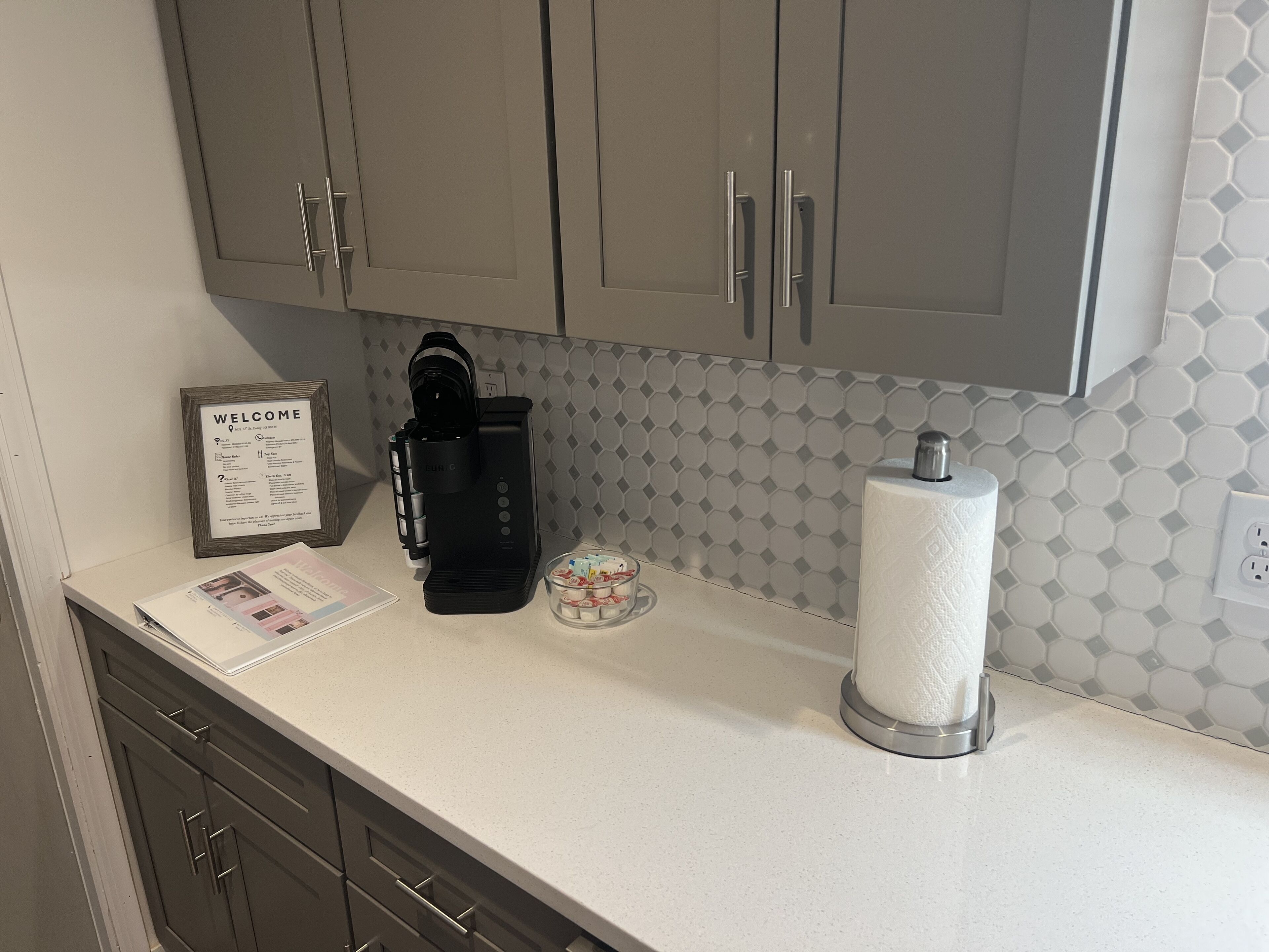 Una cocina con gabinetes grises, una cafetera, un cartel de bienvenida y un dispensador de toallas de papel.
