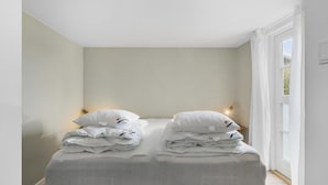 Ein ordentlich bezogenes Bett mit Kissen und gefalteten Decken, eine Nachttischlampe und ein Fenster mit Vorhängen.