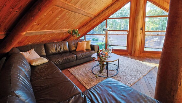 Une chambre à coucher confortable, aménagée en style « loft », avec une grande fenêtre, un plafond en bois, un canapé en cuir et une table basse.
