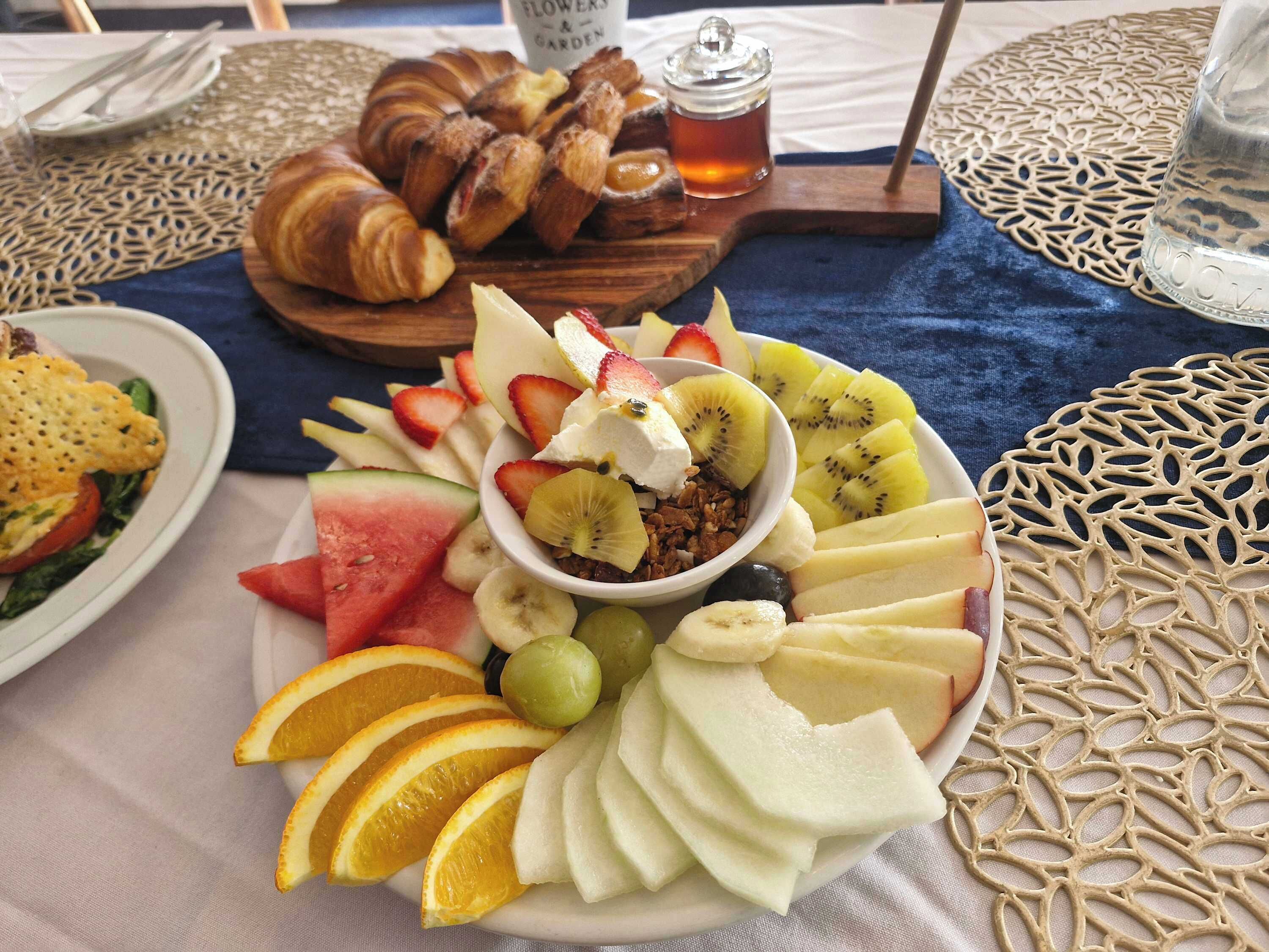 Een ontbijttafel met croissants, een schaal fruit en granola, en een glas thee op een tafelonderzetter.