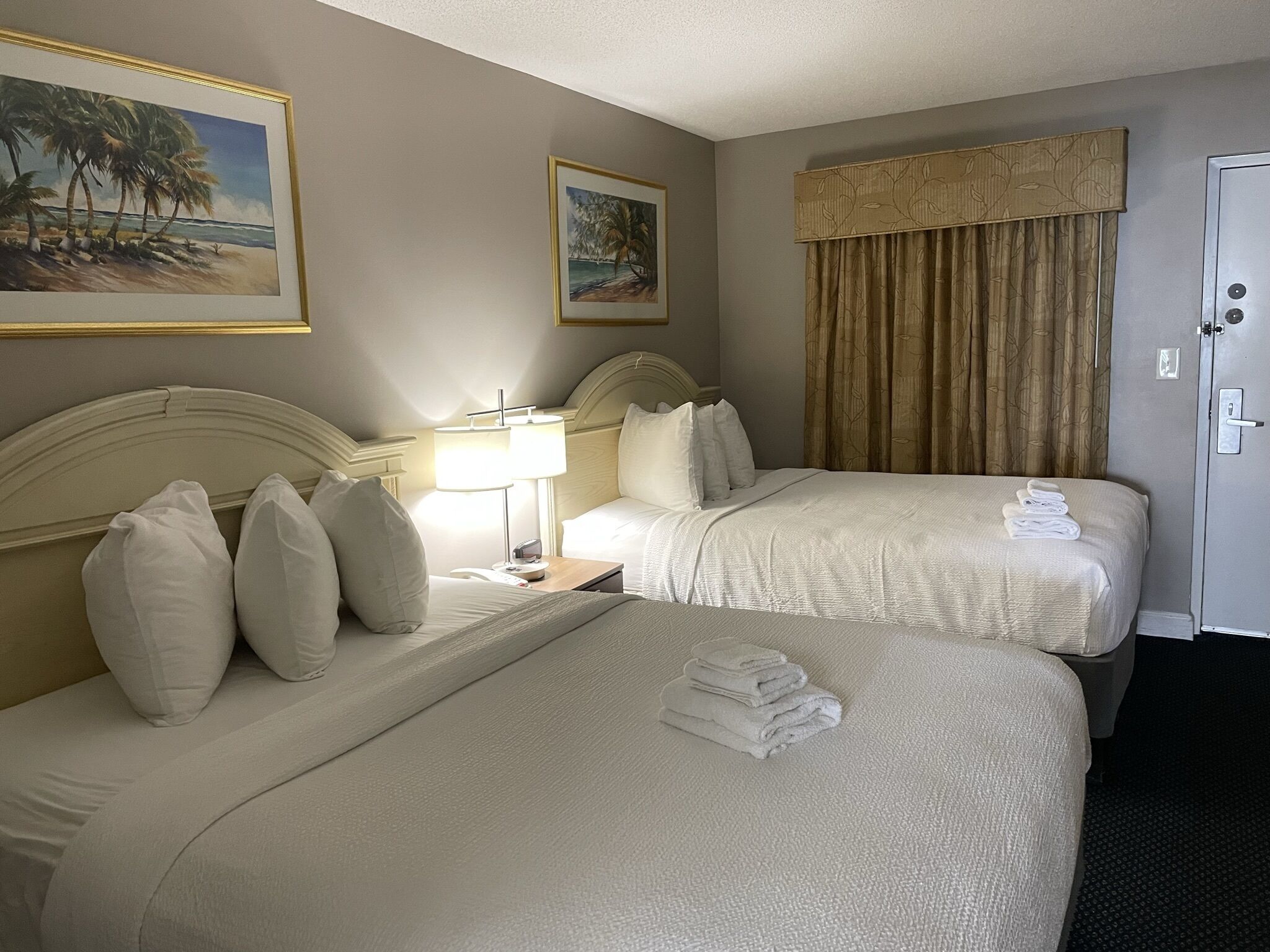 A hotel room with two beds, a nightstand with a lamp, a painting of a beach scene, and a door with a lock.