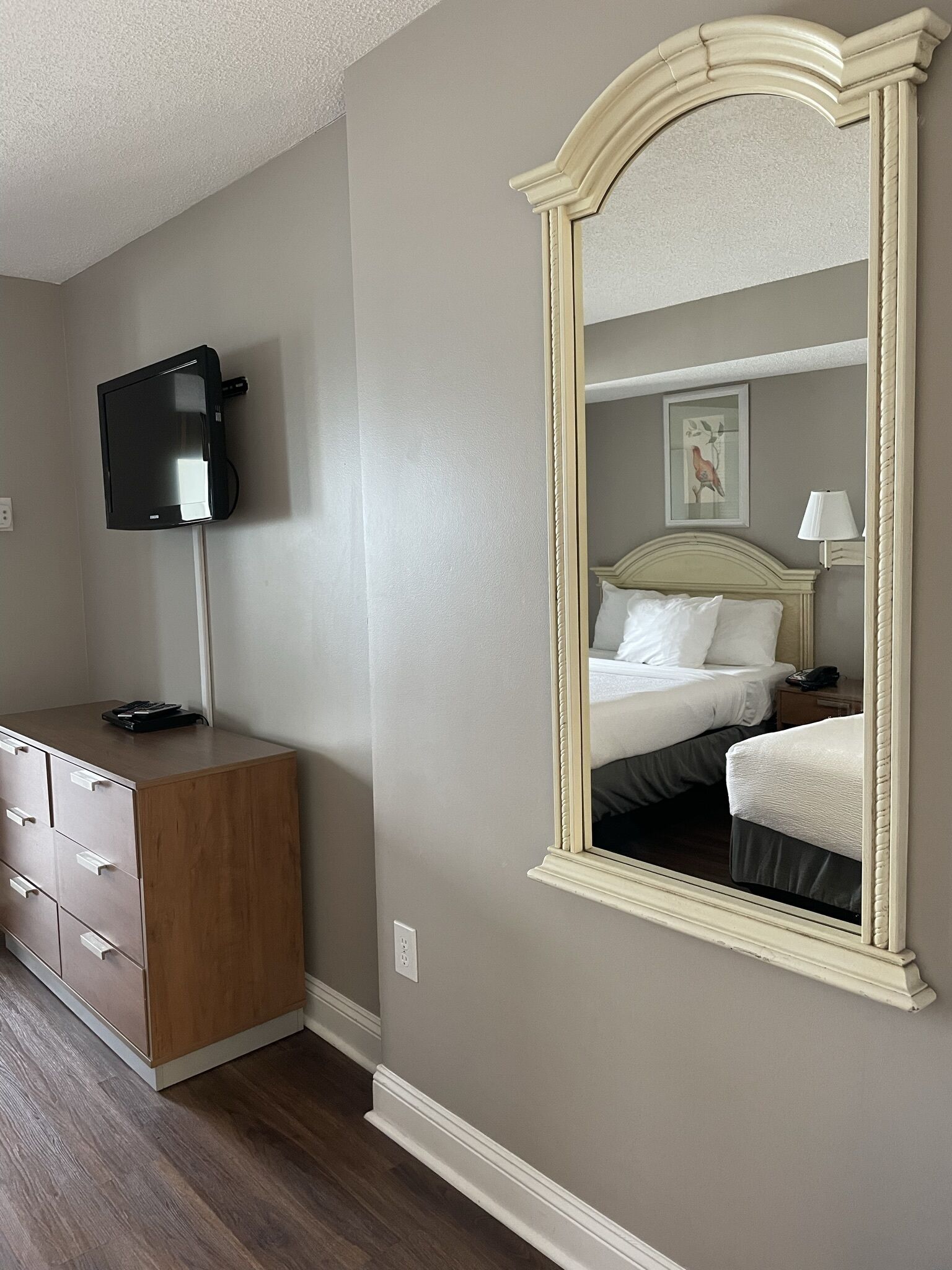 A bedroom with a large mirror, a bed, a bedside table with a lamp, and a TV mounted on the wall.