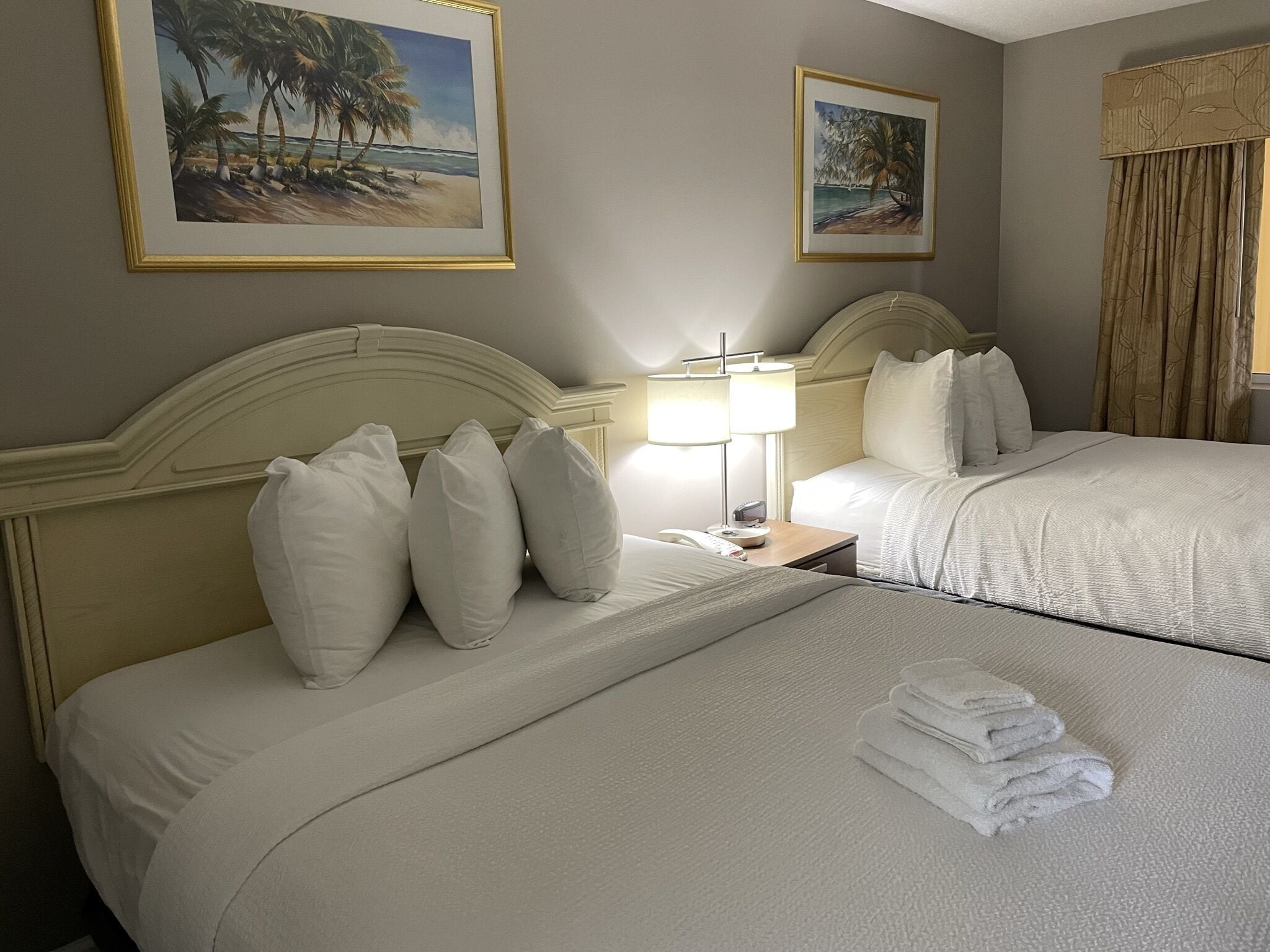 A hotel room with two beds, a nightstand with a lamp, and two framed beach paintings on the walls.