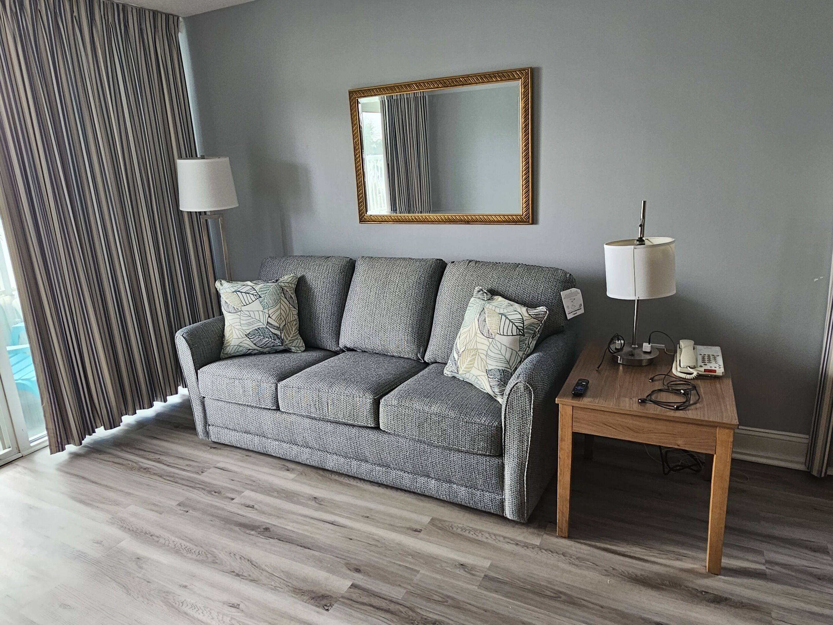 A modern living room with a grey sofa, a wooden side table, a lamp, and a mirror on the wall.