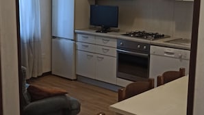 Cucina compatta con mobili bianchi, fornello, forno e frigorifero. Sono presenti un tavolo con sedie e un piccolo divano nell'angolo.