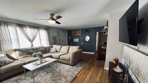 A living room with a sectional sofa, a coffee table, a TV mounted on the wall, and a ceiling fan.