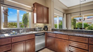 A kitchen with wooden cabinets, a stainless steel dishwasher, and a large window offering a view of greenery and mountains. - La Quinta Resort-Style Retreat (La Quinta)